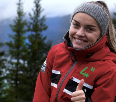 Jente i rød jakke og grått pannebånd står ute i skogen. Hun smiler og gir tommel opp til kamera. På jakka har hun en grønn 4H logo