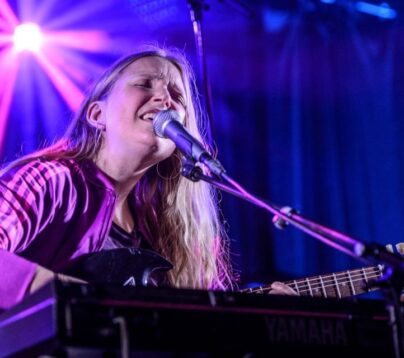 En jente sitter på en scene og synger inn i en mikrofon. Hun spiller gitar. Scenen er opplyst med lilla lyskastere. Hun er medlem i AKKS Norge