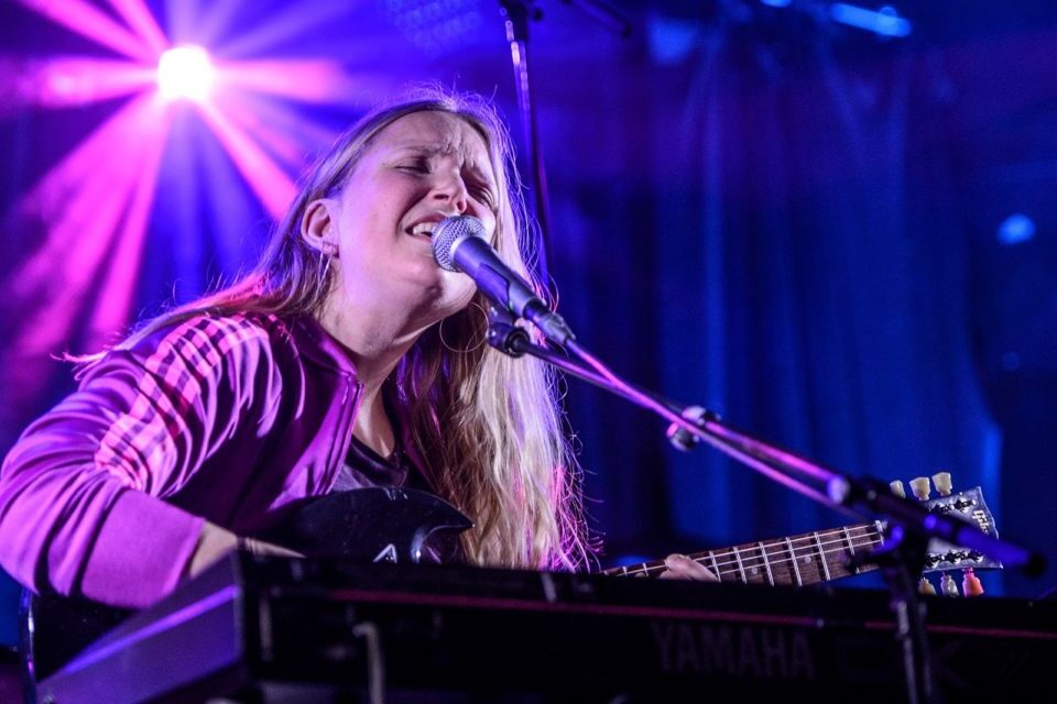 En jente sitter på en scene og synger inn i en mikrofon. Hun spiller gitar. Scenen er opplyst med lilla lyskastere. Hun er medlem i AKKS Norge