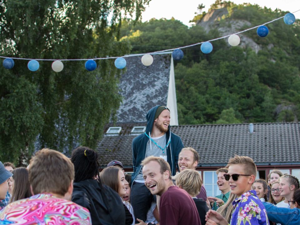 Bildet viser en forsamling mennesker som danser ute. Det er sommer, og henger vimpler i trærne. I fokus er en gutt med kort hår, briller, og hawaii-skjorte. Bildet er tatt i forbindelse med et arrangement hos NMSU.