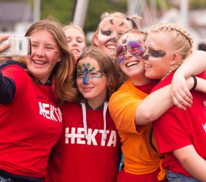 Fire jenter i Tskjorter med logoen til Hekta holder rundt hverandre og tar en selfie. Tre av dem er malt som dyr i ansiktet. De smiler og ler.