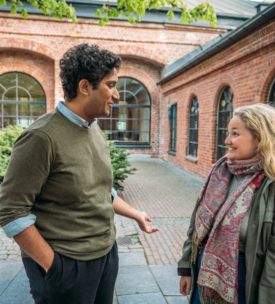 To mennesker står foran en murbygning og prater. Til venstre står det en mann med grønn genser, svarte bukser, og mørkt håt. Til høyre står det en kvinne med grønn jakke, fargerikt skjerf, og blondt hår. De smiler og prater sammen.