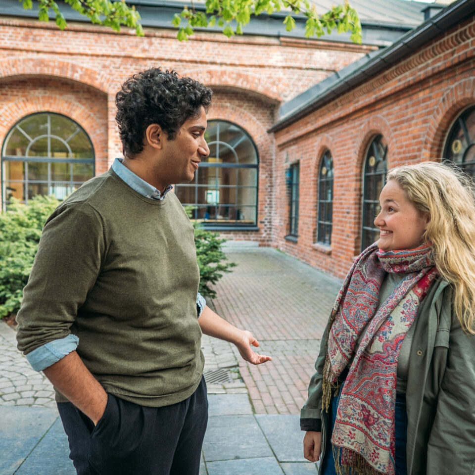 To mennesker står foran en murbygning og prater. Til venstre står det en mann med grønn genser, svarte bukser, og mørkt håt. Til høyre står det en kvinne med grønn jakke, fargerikt skjerf, og blondt hår. De smiler og prater sammen.