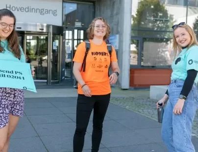 Tre unge kvinner står foran et bygg og smiler. De har på seg Tskjorter fra lærerstudentene i oransje og turkis.