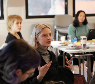 illustrasjonsbilde. Ung kvinne som stiller et spørsmål under et kurs. hun ser konsentrert ut og gestikulerer. De andre menneskene på bildet er ute av fokus, men følger tydelig med på hva kvinnen spør om.