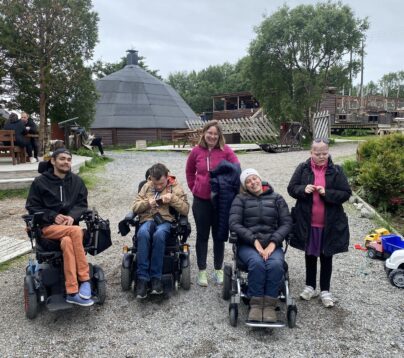 Fem mennesker på en grusvei. Tre av de sitter i rullestol. I bakgrunnen er det et stort rundt bygg, og en innhengning for hundekjøring.