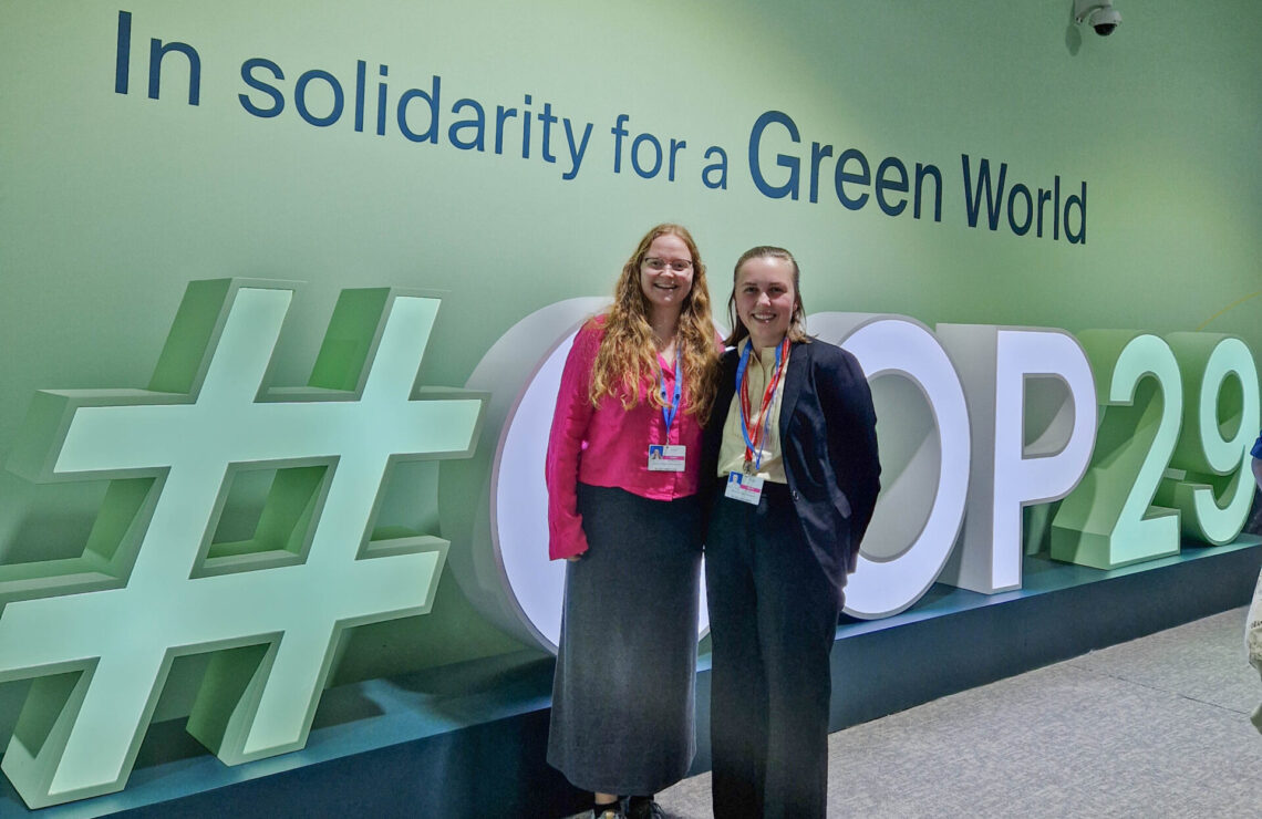 To jenter stor foran en grønn vegg. På veggen stor det In solidarity for a greener world. Det er et store bokstaver som staver #COP29 på gulvet. En av jentene har rosa bluse og langt rødt hår. Den andre har kort brunt hår og svart dress. De er ungdomsdelegater fra LNU.