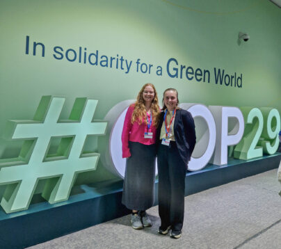 To jenter stor foran en grønn vegg. På veggen stor det In solidarity for a greener world. Det er et store bokstaver som staver #COP29 på gulvet. En av jentene har rosa bluse og langt rødt hår. Den andre har kort brunt hår og svart dress. De er ungdomsdelegater fra LNU.