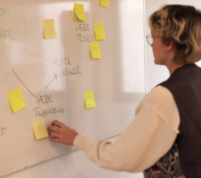 En jente med kort hår, briller, og brun vest står foran en white-board tavle. Hun er deltaker på et trygg kurs hos LNU. På tavla henger det postit lapper.