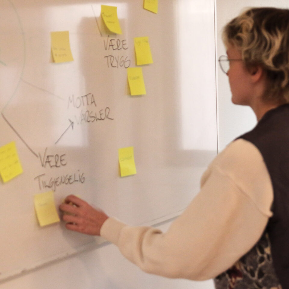 En jente med kort hår, briller, og brun vest står foran en white-board tavle. Hun er deltaker på et trygg kurs hos LNU. På tavla henger det postit lapper.