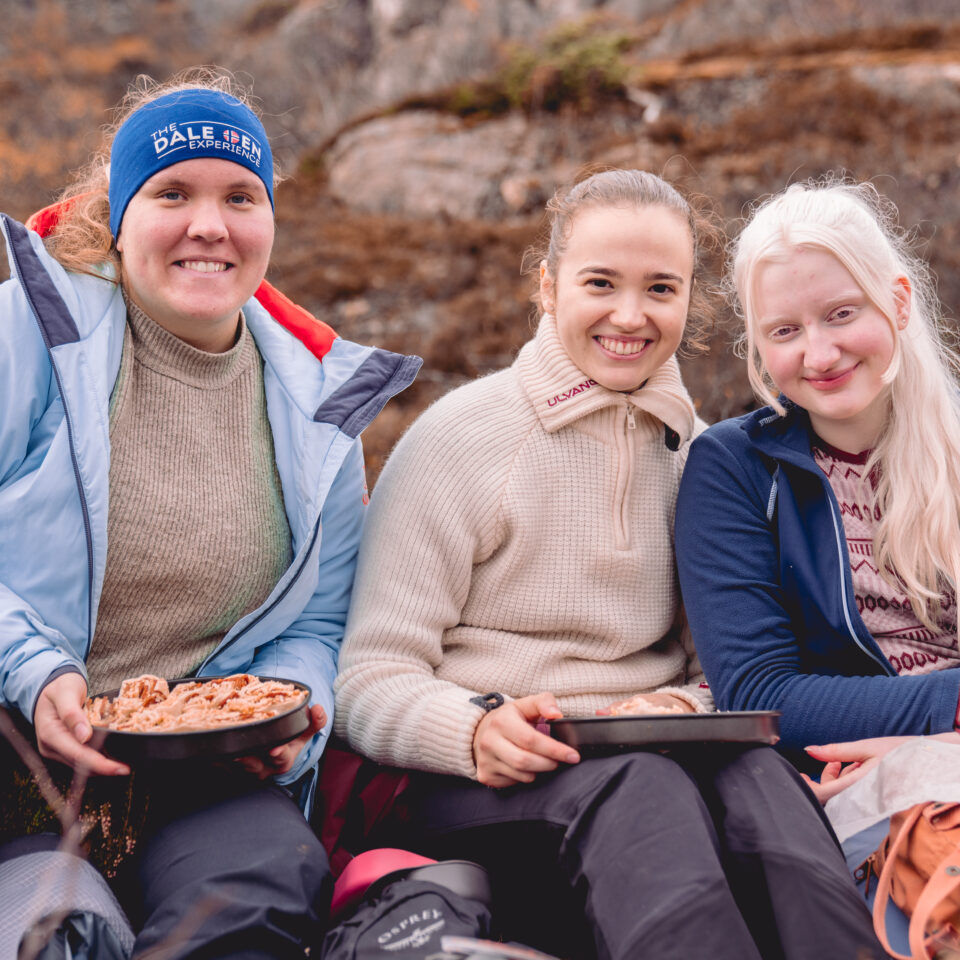 Tre damer sitter ute i naturen. De er kledd i turtøy, og holder tallerkener med turmat. De er frivillige i Norges Blindeforbunds ungdom.